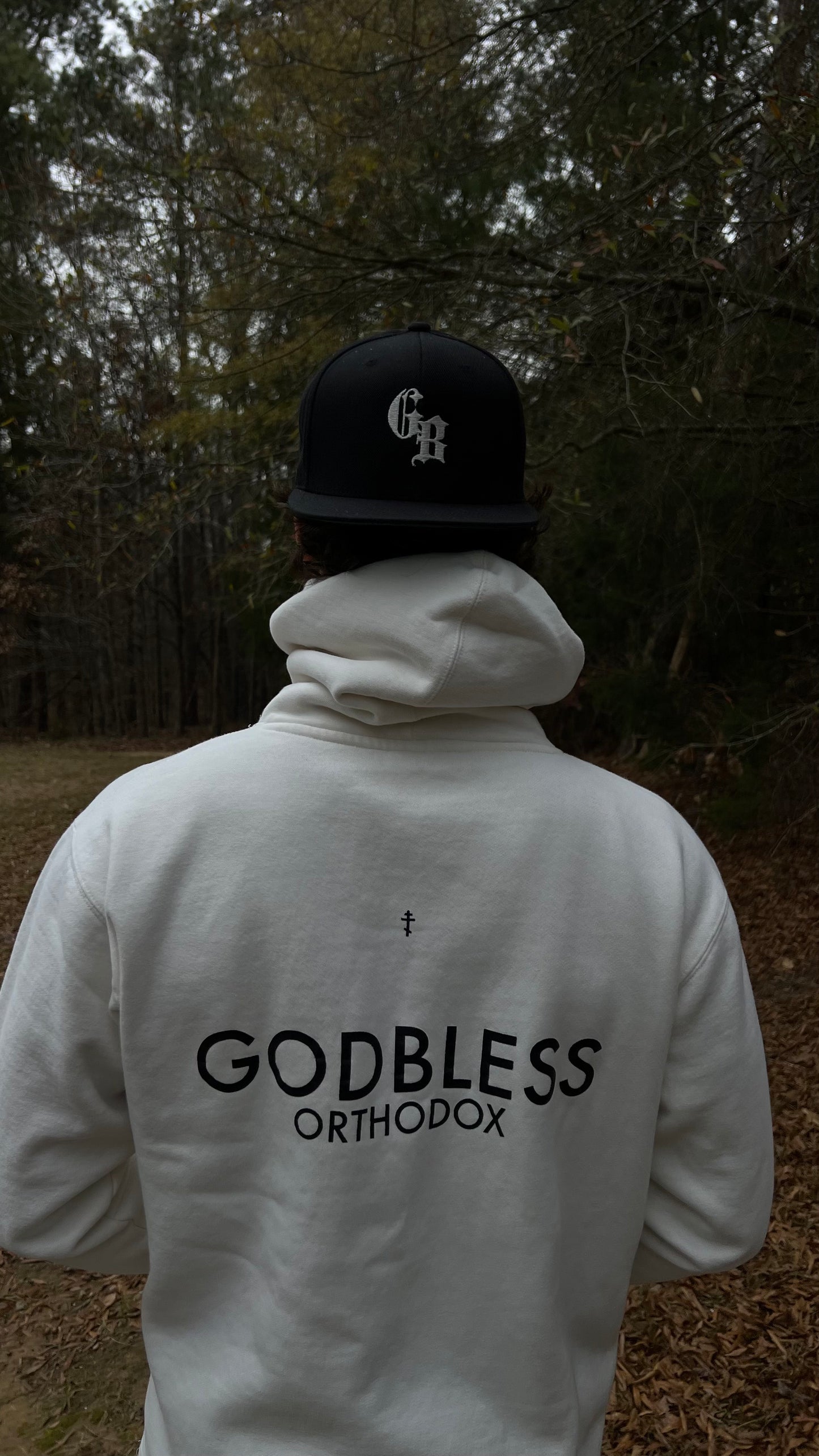 Man wearing an off-white Godbless Orthodox hoodie and a black Godbless Orthodox cap, standing in a forest.
